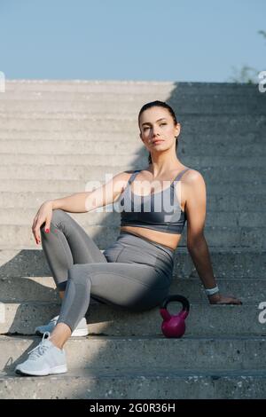 Stilvolle Brünette sitzt auf der langen Treppe im Freien, posiert mit einem niedlichen kleinen rosa Kettlebell. In grauem Oberteil und Yogahose. Am Morgen unter einer cl Stockfoto