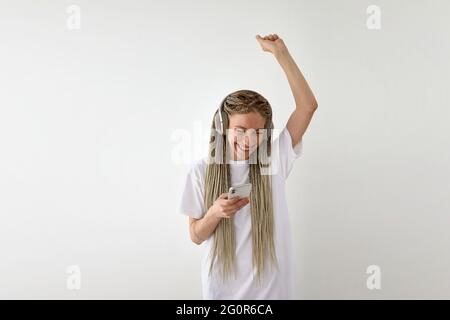 Fröhliche junge Frau im weißen Hemd, die Musik in Kopfhörern hört und mit erhobenem Arm tanzt, während sie im Studio das Smartphone auf weißem Hintergrund verwendet Stockfoto