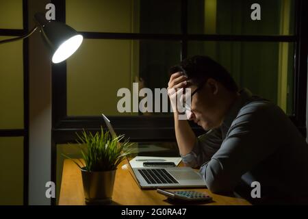 Junger asiatischer Mann, der spät in der Nacht auf dem Schreibtisch sitzt und im Büro einen Laptop anschaut, der sich ernsthaft überlegt und belastet fühlt. Überstunden und Stockfoto