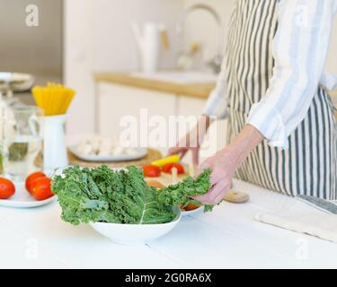 Kurzer Schuss einer pensionierten Frau auf der Schürze, die frischen gesunden Gemüsesalat zubereitet, während sie während der Pensionierung in einer modernen, hellen Küche zu Hause Lebensmittel für die ganze Familie kocht, frisches Bio-Gemüse auf dem Tisch Stockfoto