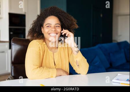 Lächelndes dunkelhäutig lockiges Mädchen hat Telefongespräch. Afrikanische Frau sitzt im Heimbüro und spricht auf dem Smartphone Stockfoto