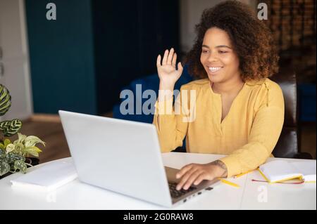 Fröhliche junge Frau nimmt am Online-Webinar, virtuellen Treffen, mit Laptop für Videoverbindung, sitzt am Schreibtisch im Heimbüro, afrikanische Frau winkt in Webcam und lächelt Stockfoto