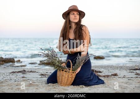 Schöne junge Frau, die einen Korb mit wilden Blumen in der Hand hält und am Strand sitzt. Weichfokus Stockfoto