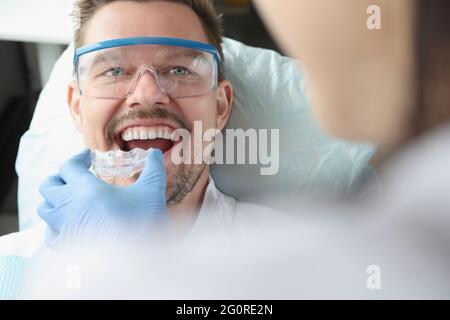 Der Arzt versucht, einen Mundschutz aus Kunststoff zu nutzen, damit ein junger lächelnder Mann den Bissen korrigieren kann Stockfoto