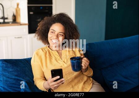 Eine ruhige, dunkelhäutige, afroamerikanische Frau verbringt das Wochenende zu Hause, sitzt mit einem Becher in den Händen auf dem Sofa, nutzt das Smartphone, genießt Kaffee und chattet online Stockfoto