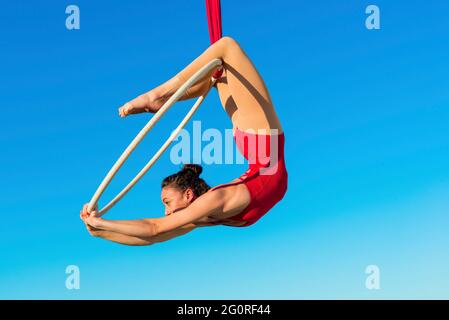 acrobat athletische, junge anmutige Turnerin Durchführung Luftübungen im Luftring im Freien auf Himmel Hintergrund. Flexible Frau in rotem Anzug führt Stockfoto