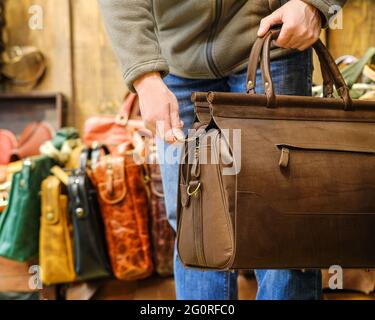 Die Hand eines Mannes, der eine braune Reisetasche vor dem Hintergrund anderer Taschen im Laden hält. Das Konzept der Naturlederprodukte Stockfoto