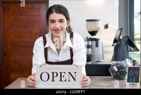 Der junge kaukasische Ladenbesitzer hält mit einem Lächeln ein OFFENES Schild vor einem Coffee-Shop-Schalter. Morgendliche Atmosphäre in einem Café. Stockfoto