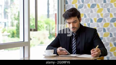 Kaukasischer Geschäftsmann in schwarzem Anzug, der während der Mittagspause eine Notiz auf dem Mobiltelefon liest. Atmosphäre in einem modernen Café. Stockfoto