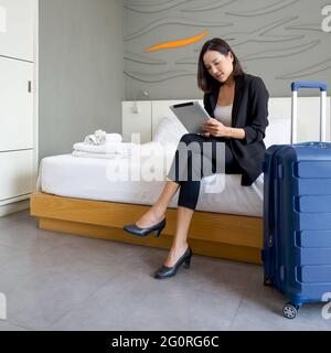 Asiatische Geschäftsfrau in schwarzem Anzug sitzt mit Tablet-Computer in der Hand. Ihr Gepäck wurde neben das Bett gelegt. Die Atmosphäre der Arbeit im Freien während des Th Stockfoto