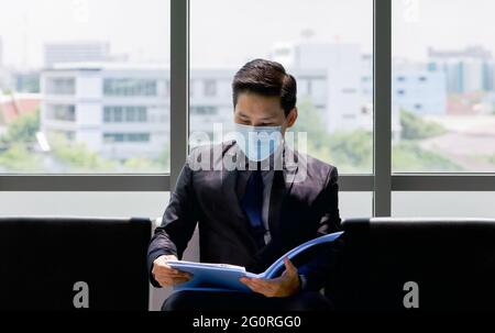 Asiatischer Geschäftsmann in Anzug und Gesichtsmaske sitzt auf einer Bank in der Nähe des Fensters, untersucht seinen Lebenslauf, während er auf das Interview wartet. Stockfoto