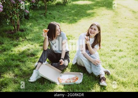 Zwei junge Teenager-Freunde, die sich im Park auf dem Rasen amüsieren und Pizza essen. Frauen essen Fast Food. Keine gesunde Ernährung. Weicher, selektiver Fokus. Stockfoto