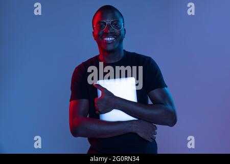 Glücklicher junger afrikanischer hübscher Mann mit Tablet isoliert über dunklem Hintergrund in gemischtem Neonlicht. Stockfoto
