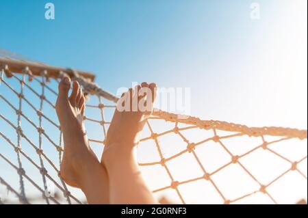 Blick auf die Frau, die sich in der Hängematte entspannt. Low-Angle-Ansicht. Wunderschöne weibliche Füße, die sich in einer Hängematte am Strand vor dem Hintergrund des Himmels entspannen Stockfoto