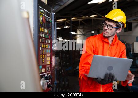 Leitender Ingenieur, der in der Fabrik einen Hut trägt und einen Laptop in der Hand hält, hübscher Arbeiter in schweren Industrieumgebungen Stockfoto