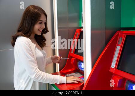 Junge asiatische Frau, die Geld mit einer Karte am Automaten abzieht, Frau, die am Geldautomaten der Bank steht Stockfoto