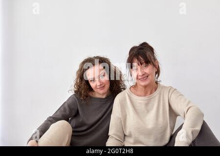 Fröhliche Erwachsene Tochter und Mutter mittleren Alters sitzen auf dem Boden zu Hause auf weißem Hintergrund Stockfoto