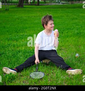 Ein Junge isst Eis auf dem Rasen im Park mit einem Badmintonschläger Stockfoto