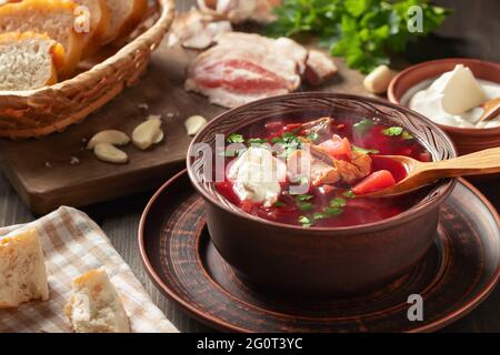 Frisch zubereitetes, warmes, hausgemachtes Borscht: Traditionelles Gericht der russischen und ukrainischen Küche in Steingutgerichten mit Speck, Brot, Sauerrahm und Knoblauch Stockfoto