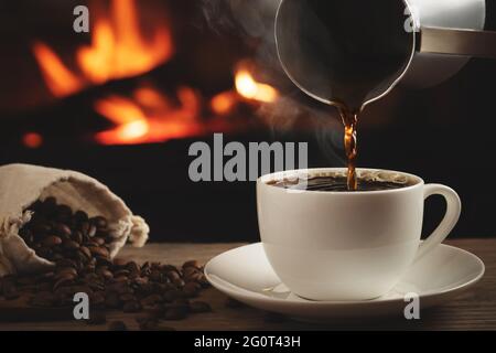 Gießen von Cezve-Kaffee in eine Tasse auf einem Holztisch vor einem brennenden Kamin. Selektiver Fokus. Stockfoto