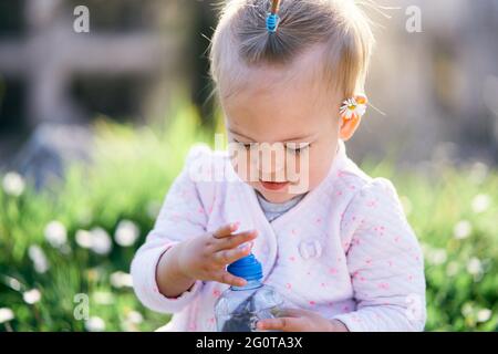 Das kleine Baby mit einer Kamille hinter ihrem Ohr sitzt auf einem grünen Rasen und öffnet eine Plastikflasche. Nahaufnahme Stockfoto