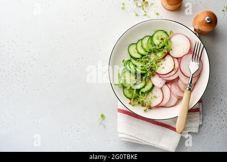 Frischer Salat mit rotem Rettich, Gurke, Gemüse, mikrogrünen Radieschen auf weißem Teller auf grauem Steingrund. Blick von oben. Konzept vegan und hea Stockfoto