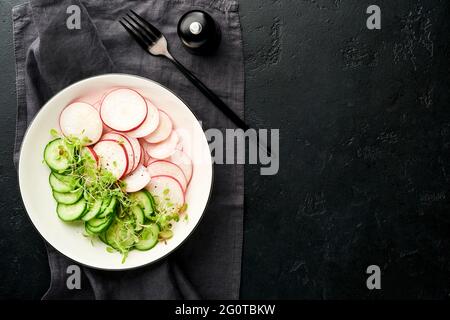 Frischer Salat mit rotem Rettich, Gurke, Gemüse, mikrogrünen Radieschen auf weißem Teller auf grauem Steingrund. Blick von oben. Konzept vegan und hea Stockfoto