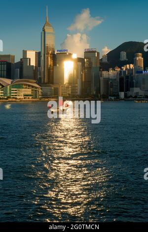 Die Aqua Luna, eine nachgebaute chinesische Dschunke, die für Sightseeing-Kreuzfahrten verwendet wird, durchläuft die Spiegelung der späten Nachmittagssonne vom Hochhaus ... Stockfoto