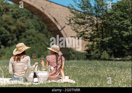 Zwei junge Frauen mit Strohhut sitzen auf einer Decke in einem Garten voller Blumen. Weibchen halten ein Glas Weißwein in der Hand und sehen. Stockfoto