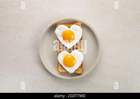 Herzförmiges Ei in getoasteten Roggenbrotscheiben auf Keramikplatte. Ich liebe das Frühstücksdesign. Gesundes Sandwich. Festliches Mittagessen oder Frühstück. Eiertag. Stockfoto