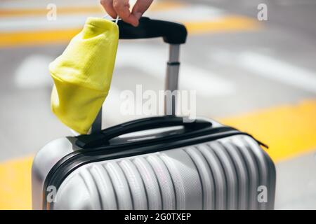 Hand hält grauen Reisegepäckkoffer und gelbe Schutzmaske, Straße an einem sonnigen Tag. Covid und sicheres Reisekonzept Hintergrund für Textkopie Stockfoto