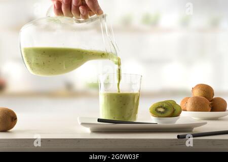 Person, die Kiwi trinkt, aus einem Krug mit geschnittenem Obst in Behältern auf der Küchenbank. Vorderansicht. Stockfoto