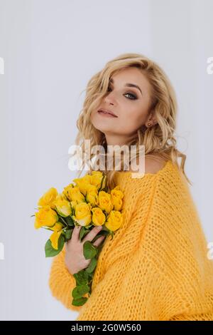 Porträt einer schönen jungen blonden Frau mit einem Strauß gelber Rosen in einem voluminösen Pullover auf weißem Hintergrund im Studio. Weich selektiv Stockfoto