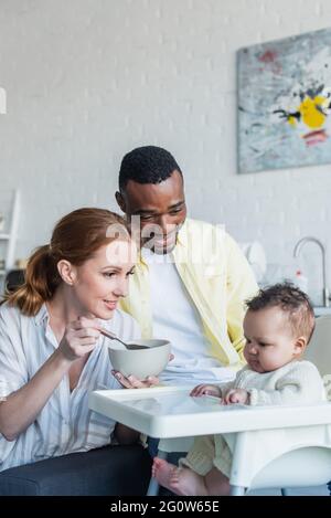 Glückliche Frau Fütterung Kleinkind in der Nähe afroamerikanischen Ehemann Stockfoto