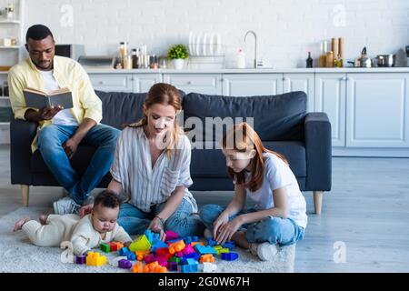 Glückliche Frau spielt mit interracial Töchtern in der Nähe afroamerikanischen Ehemann Buch auf der Couch lesen Stockfoto