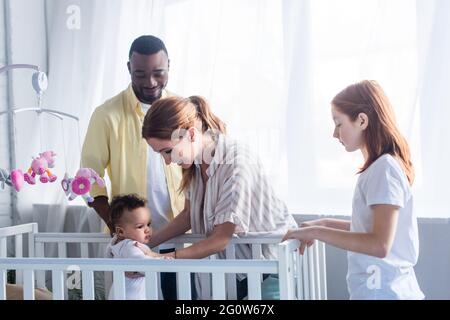 Glückliche Frau unterstützt Baby Mädchen in der Krippe in der Nähe multiethnischen Familie stehen Stockfoto