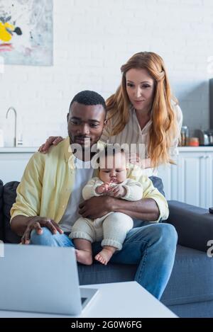 Multiethnisches Paar mit Kleinkind, die auf der Couch in der Nähe des Laptops sitzt Stockfoto