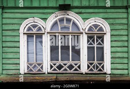 Dreifach gewölbtes Fenster mit weißem Holzrahmen in grungigen grünen Holzwand, russische ländliche Architektur Hintergrund Fotofragment Stockfoto