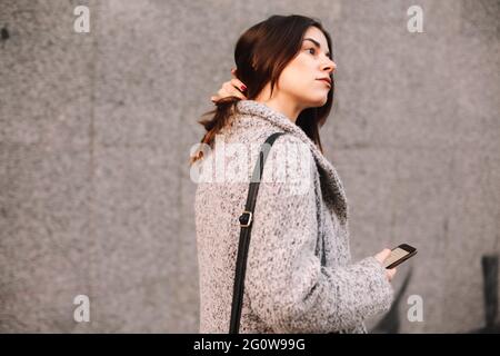 Nachdenkliche Frau mit Smartphone, die gegen die graue Wand in der Stadt läuft Stockfoto