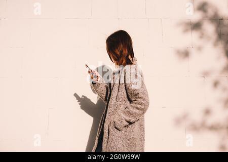 Seitenansicht einer nicht-binären Frau, die ein Smartphone gegen die Wand benutzt Stockfoto