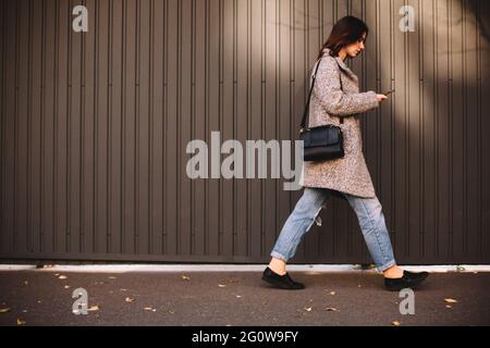 Seitenansicht einer nicht-binären Frau, die ein Smartphone benutzt, das gegen die Wand läuft Stockfoto