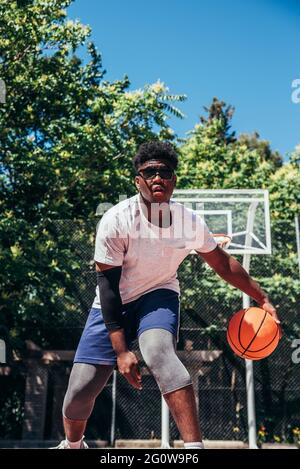 Schwarzer afroamerikanischer Junge mit Sonnenbrille, der auf einem städtischen Platz Basketball spielt. Stockfoto