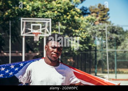 Porträt eines schwarzen afroamerikanischen Jungen, der die US-Flagge auf einem städtischen Basketballplatz trägt. Stockfoto