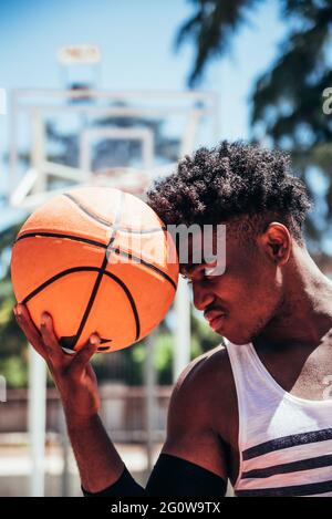 Porträt eines schwarzen afroamerikanischen Jungen, der einen Basketball in der Stirn auf einem städtischen Basketballplatz hält.