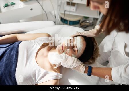 Hochwinkelansicht eines Ästhetikers, der Schönheitsbehandlungen auf dem Gesicht einer Frau durchführt, wobei der Rest des kosmetischen Produkts mit speziellen Nassservietten entfernt wird. Bekennen Stockfoto