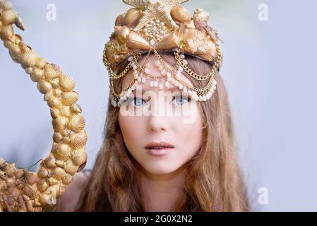 Fantasy Frau echte Meerjungfrau mit Dreizack Mythos Göttin des Meeres mit goldenen Schwanz sitzt in Sonnenuntergang auf Felsen.. Gold Haar Krone Muscheln Perlen Schmuck. Merma Stockfoto