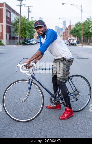 Junger Afroamerikaner mit Fahrrad, das draußen auf der Straße mit städtischem Hintergrund in Chicago, IL, USA steht. Stockfoto