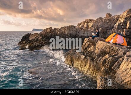 Frau, die auf dem Felsen über dem Meer sitzt und am Computer arbeitet. Geschäftsfrau, die vom Strand aus arbeitet. „Arbeiten aus der Ferne“-Konzept. Freiberufler tra Stockfoto