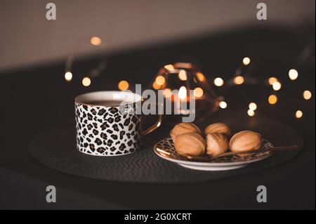 Frische Tasse Tee mit leckerem Gebäck auf dem Teller mit brennender Kerze und Weihnachtsbeleuchtung aus der Nähe. Winterferien. Gemütliche Atmosphäre. Feiern Stockfoto