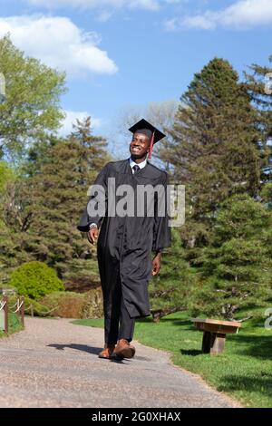 Ein junger, schwarzer Hochschulabsolvent mit Mütze und Kleid lächelt, als er stolz einen Pfad entlang geht Stockfoto